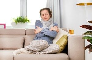 Woman on couch looking cold due to furnace malfunctioning. Beige couch, woman wearing gray sweater and gray scarf.